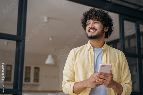 Handsome smiling Indian man holding mobile phone shopping online looking away at home, copy space  photo