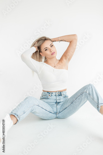 Young beautiful blonde girl in a fashionable white T-shirt with blue vintage jeans sits on a white background in the studio