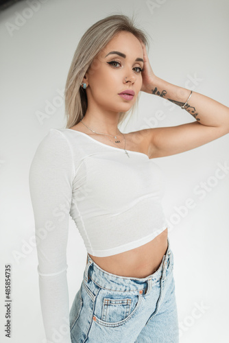 Fashion cute girl with blonde hairstyle in stylish white t-shirt and blue jeans stands and looks at the camera on a white background in studio