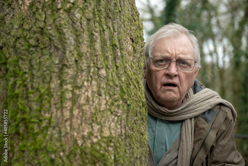 Portrait of shocked senior man peeking from behind a large tree in the woods