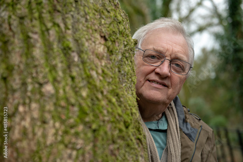 Portrait of happy senior man peeking from behind a large tree in the woods