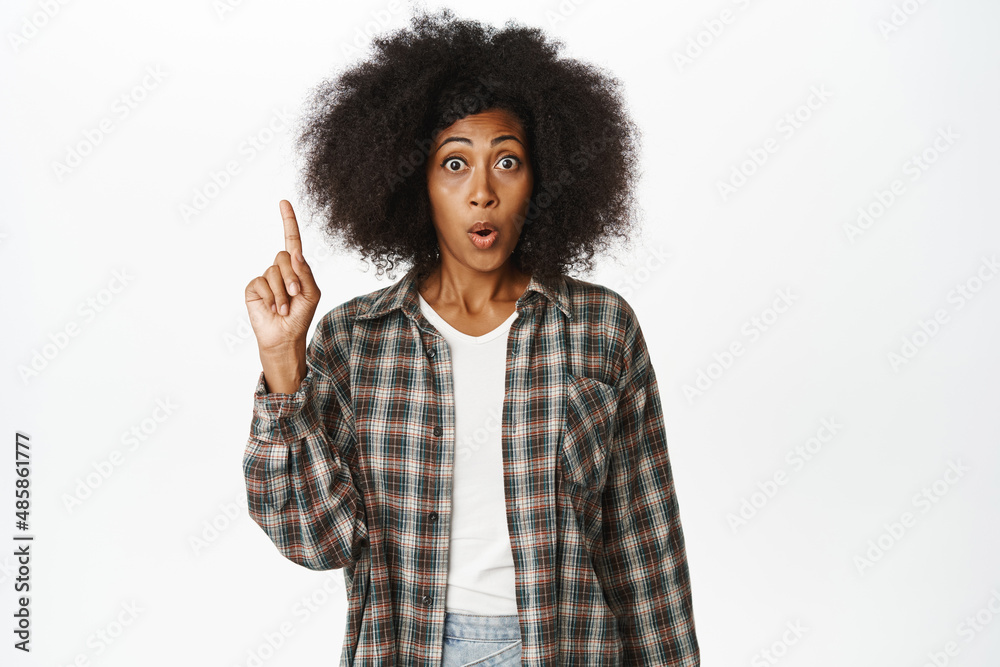 Enthusiastic african american woman pointing finger up, looking surprised and intrigued, showing advertisement, standing in casual clothes over white background Enthusiastic african american woman pointing finger up, looking surprised and intrigued, showing advertisement, standing in casual clothes over white background