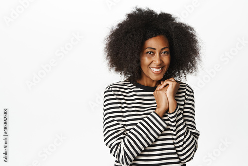 Image of cute african woman looking with love, tenderness and care, holding hands near heart, appreciate smth, standing over white background