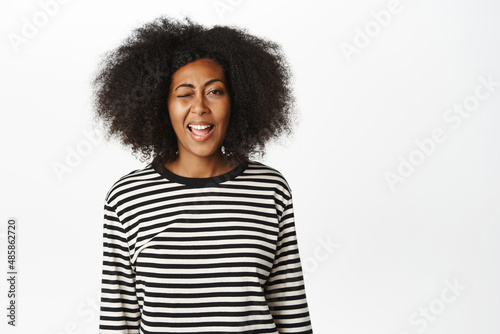 Positive Black woman in casual clothes, shows tongue, winks and smiles, expresses happy and carefree emotion, stands over white background