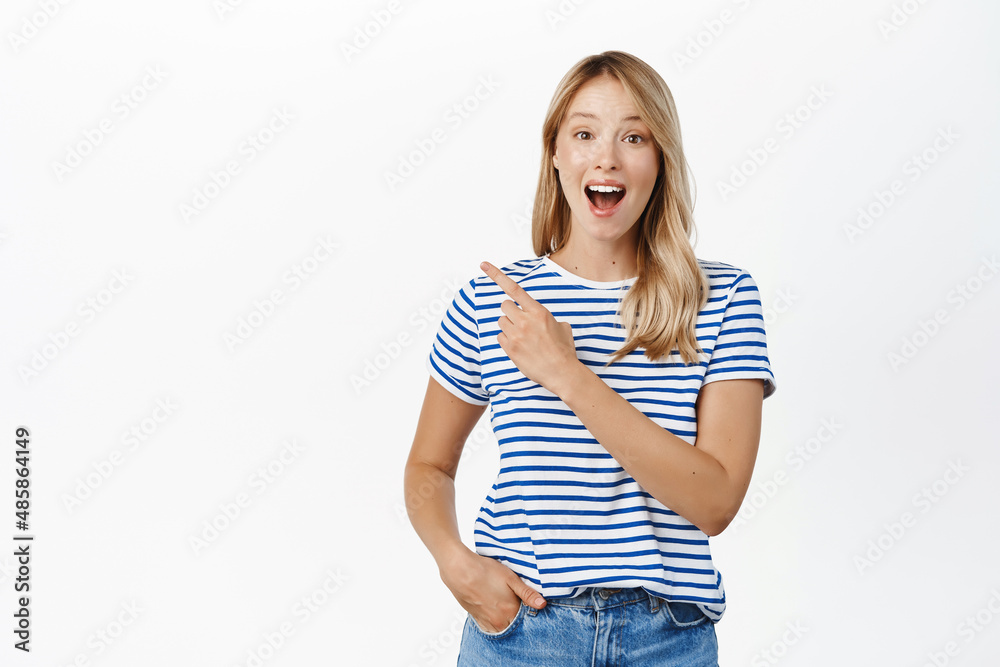 Happy smiling blond woman pointing finger left, showing announcement or banner, announcing event, standing in tshirt over white background Happy smiling blond woman pointing finger left, showing announcement or banner, announcing event, standing in tshirt over white background