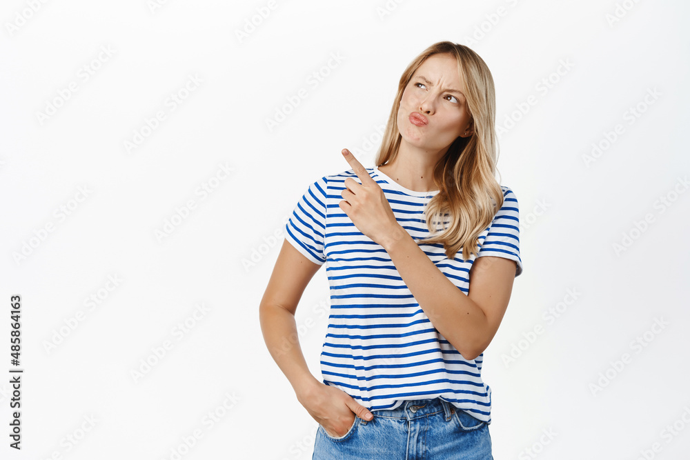 Portrait of young woman looks thoughtful and points at upper left empty space, promo banner on top, thinking or choosing in store, standing over white background Portrait of young woman looks thoughtful and points at upper left empty space, promo banner on top, thinking or choosing in store, standing over white background
