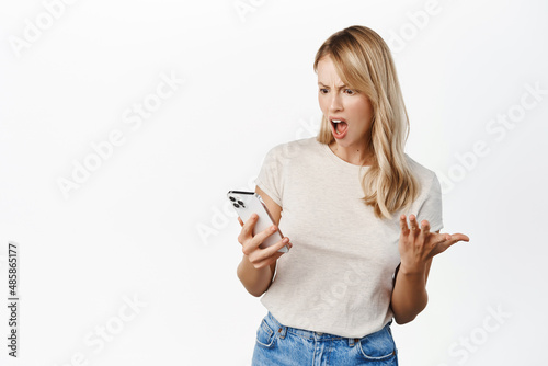 Angry young woman screaming, complaining at mobile phone, looking frustrated at cellphone, reading smth upsetting on smartphone, white background