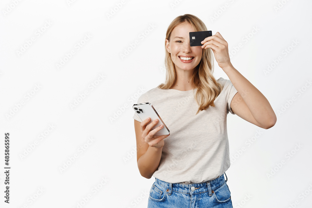 Happy smiling woman with mobile phone, showing credit card and laughing, demonstrating paying app, bank offer for contactless online shopping pay, white background Happy smiling woman with mobile phone, showing credit card and laughing, demonstrating paying app, bank offer for contactless online shopping pay, white background