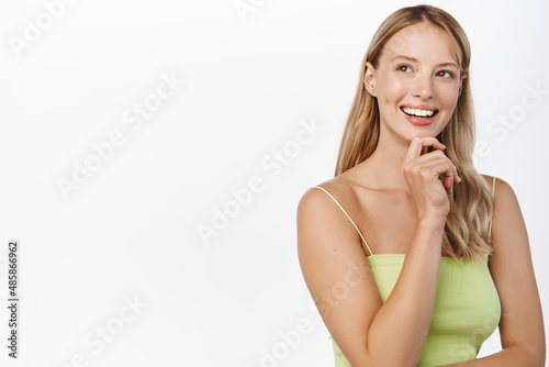 Stylish modern blond girl looking pleased at promo text, advertisement on copy space, smiling and thinking, standing over white background