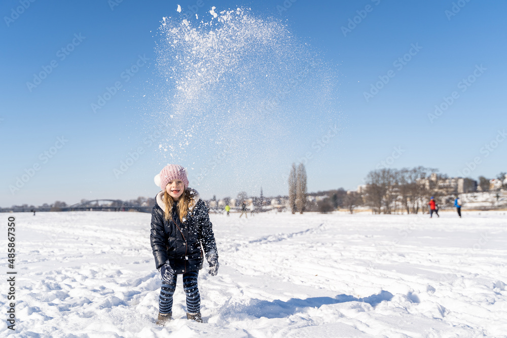 Young caucasian smiling preschool girl in warm clothes throws snow up playing and having fun on sunny winter weather outside on snow covered field in city park. Christmas holidays, childhood, carefree