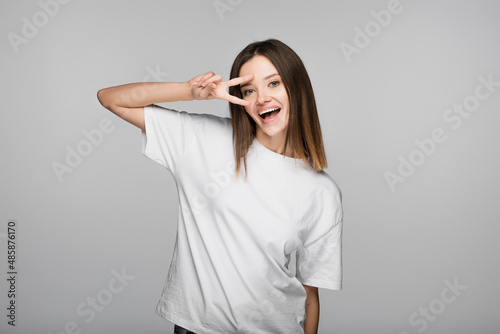 excited brunette woman showing victory sign near face isolated on grey