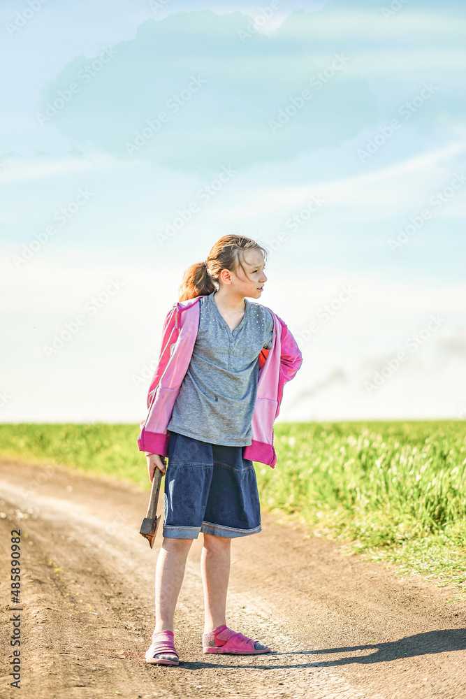 A girl on the road near the field holds an ax in her hand and looks to the side. A girl on the road near the field holds an ax in her hand and looks to the side.