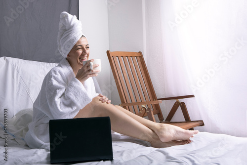 portrait of a middle-aged woman, sitting on the bed in a white bathrobe after a bath, with a large white cup while laughing