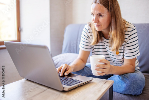 Young attractive woman using laptop computer and drinking morning cup of coffee while wearing a jeans and t-shirt. Brunette female sitting in a living room. Freelancer working from home