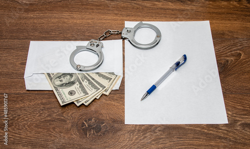 Handcuffs, an envelope with dollars, a pen and a blank sheet of paper on a wooden table. Frank confession, arrest, interrogation. Punishment for illegal income, bribe.