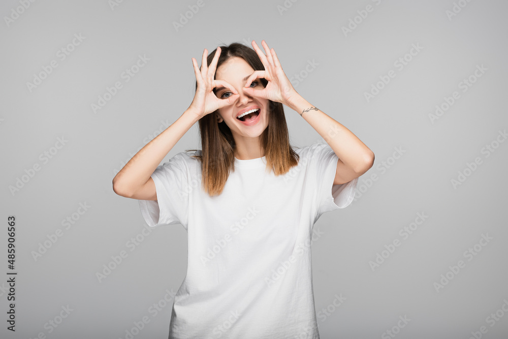 cheerful woman in white t-shirt showing okay signs near eyes isolated on grey cheerful woman in white t-shirt showing okay signs near eyes isolated on grey
