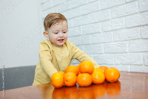 A little boy 2 years old holds tangerines in his hands. The kid wants to sit on citrus fruits for the first time.