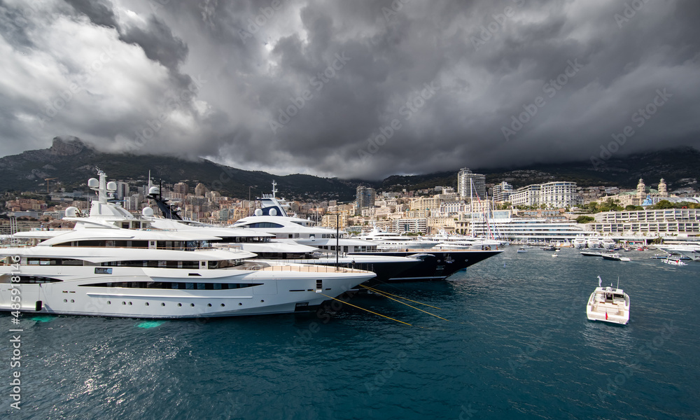 A lot of huge yachts are in port of Monaco at storm weather, mountain is on background, glossy board of the motor boat, megayachts are moored in marina, sun reflection on glossy board A lot of huge yachts are in port of Monaco at storm weather, mountain is on background, glossy board of the motor boat, megayachts are moored in marina, sun reflection on glossy board