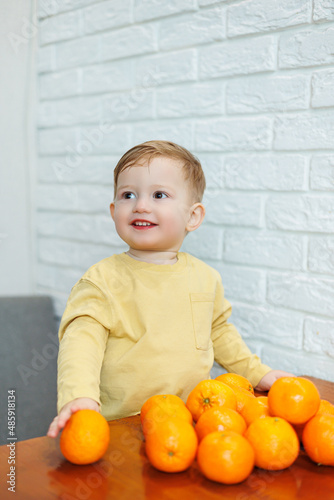 A little boy 2 years old holds tangerines in his hands. The kid wants to sit on citrus fruits for the first time.