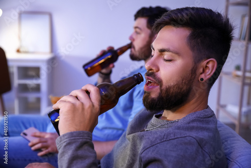 Young bearded man drinking beer at home in evening, closeup