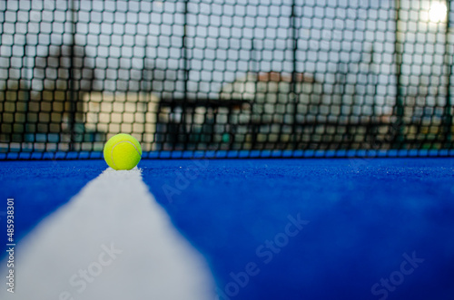 Selective focus. Ground level view of a ball on the line of a blue paddle tennis court with the net out of focus in the background.