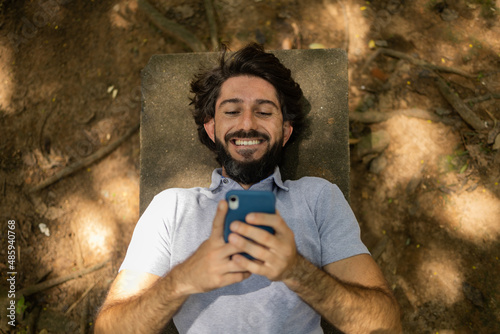 View of young man using a smartphone at day time lying down on a bench at a park. High quality photo