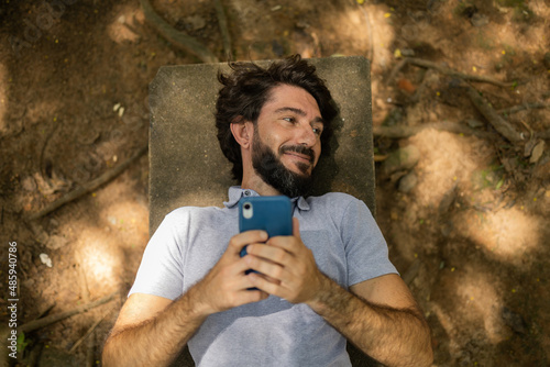 View of young man using a smartphone at day time lying down on a bench at a park. High quality photo