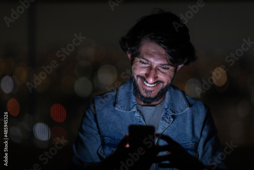 Front view of young man using a smartphone at night time with city view landscape in the background. High quality photo