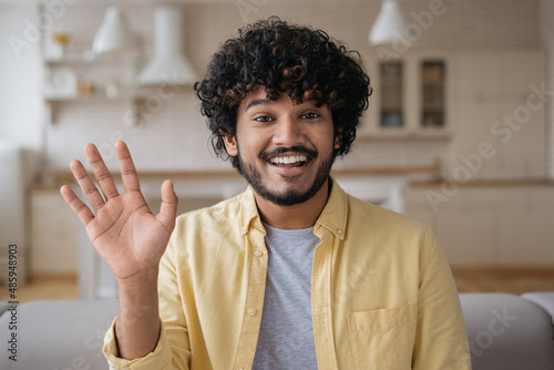 Smiling Indian man freelancer having video call working online from home. Young happy influencer recording video waving hand talking to camera photo