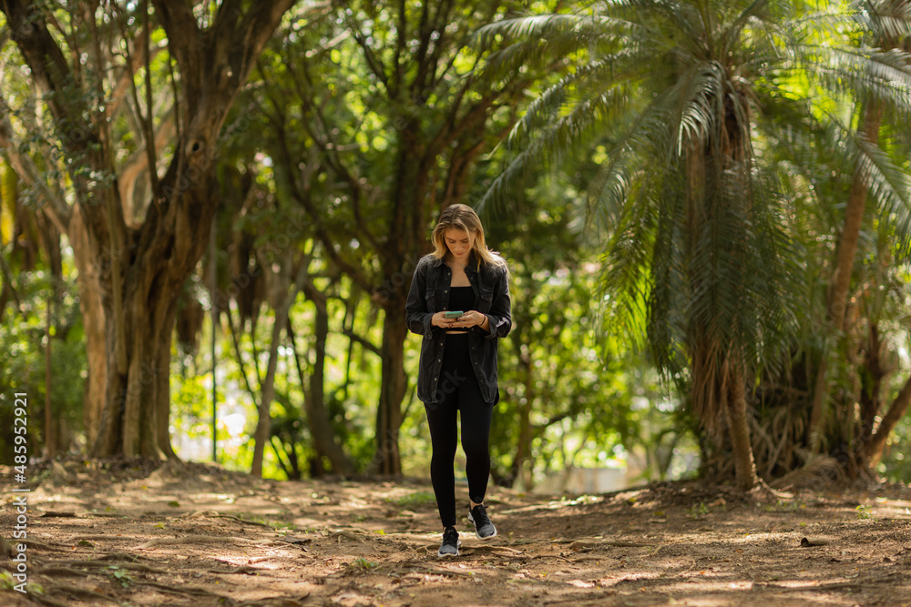 Fototapeta premium Young woman using a smartphone at day time with a green park in the background. High quality photo