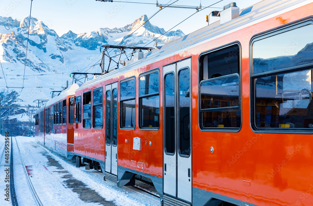 Modern red train running on rack railway through snowy Swiss Alps on sunny winter day..