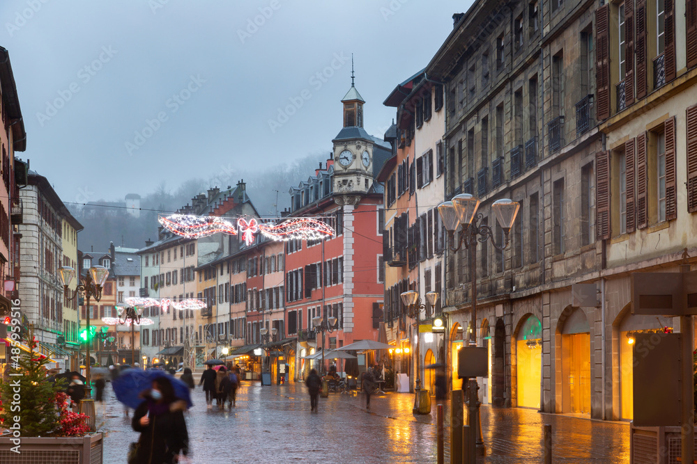 View of Place Saint-Leger, main pedestrian street in historic center of French city of Chambery decorated with traditional Christmas lights overlooking clock tower on rainy winter evening.