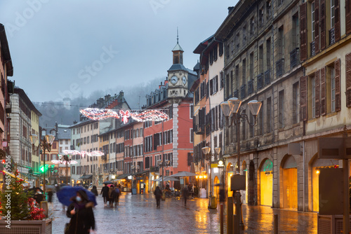 Fototapeta Naklejka Na Ścianę i Meble -  View of Place Saint-Leger, main pedestrian street in historic center of French city of Chambery decorated with traditional Christmas lights overlooking clock tower on rainy winter evening.