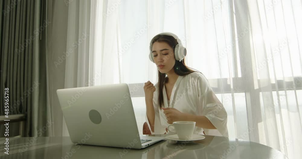 Joyful asian gen z girl wearing headphones and dancing in early morning. Positive teen having fun while using laptop computer 4k footage Joyful asian gen z girl wearing headphones and dancing in early morning. Positive teen having fun while using laptop computer 4k footage