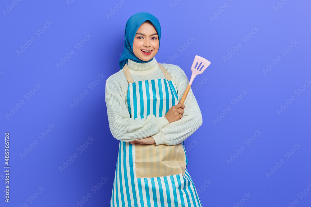 Smiling beautiful Asian woman wearing apron and headscarf holding spatula with crossed arms isolated over purple background. Housekeeping concept