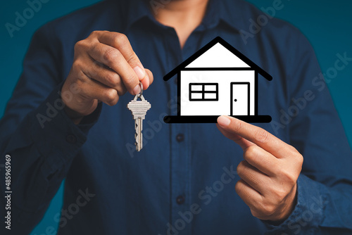 Business and real estate concept. Hand holding a mini paper house and keys while standing on a blue background.