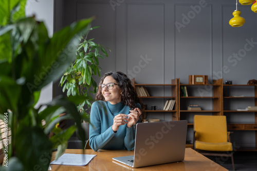 Happy female employee looking in window with satisfied face expression while sitting in office in front of laptop, woman taking break from computer work at workplace. Job satisfaction concept photo