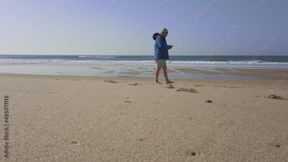 Caucasian male is walking on beach and using mobile phone, autumn weather and clothes Caucasian male is walking on beach and using mobile phone, autumn weather and clothes