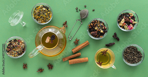 several types of herbal tea mixtures in a glass dish next to a kettle and a cup of hot tea on a green background. View from above. Banner, Healing warming aromatic drink.