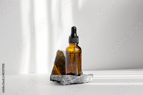 Fototapeta Naklejka Na Ścianę i Meble -  Serum in glass bottle with dropper lid on stand made of natural granite stone. Essential oil for care of women's skin on white background, in rays of sunlight. Space for copying, glare on object