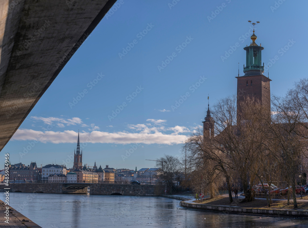 Walk way under a traffic route at the canal Karlbergskanalen, dividing modern buildings from old classic houses with the Town City Hall at the bridge Stadshusbron a sunny winter day in Stockholm