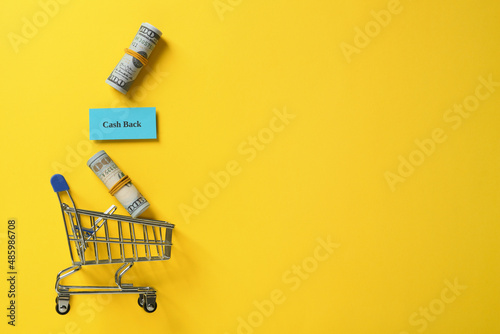 Card with word Cashback, rolled dollar banknotes and shopping cart on yellow background, flat lay. Space for text