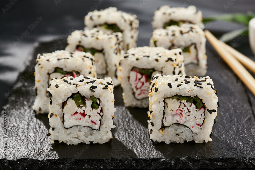Maki sushi on dark slate. California maki with sesame. Sushi roll with crab, cucumber inside, sesame outside. Style concept japanese menu with black background, leaves and hard shadow. Maki sushi on dark slate. California maki with sesame. Sushi roll with crab, cucumber inside, sesame outside. Style concept japanese menu with black background, leaves and hard shadow.
