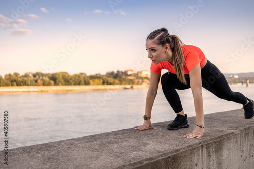 Young happy focused fitness girl in black yoga pants and orange short shirt work out and stretch her body on short concrete wall near river during the day.