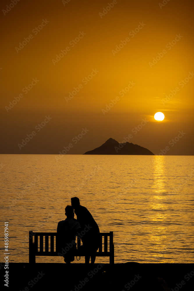 Couple relaxing on bench by sea during sunset