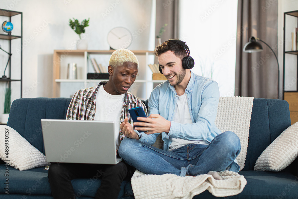 Caucasian man in headphones showing something on smartphone to his african boyfriend that sitting near with laptop on knees. Concept of relations, people and technology. Caucasian man in headphones showing something on smartphone to his african boyfriend that sitting near with laptop on knees. Concept of relations, people and technology.