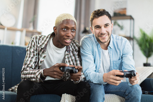 Multi ethnic same sex couple sitting together on couch and using wireless joysticks for playing video games. Happy young man spending free time time with fun at home.