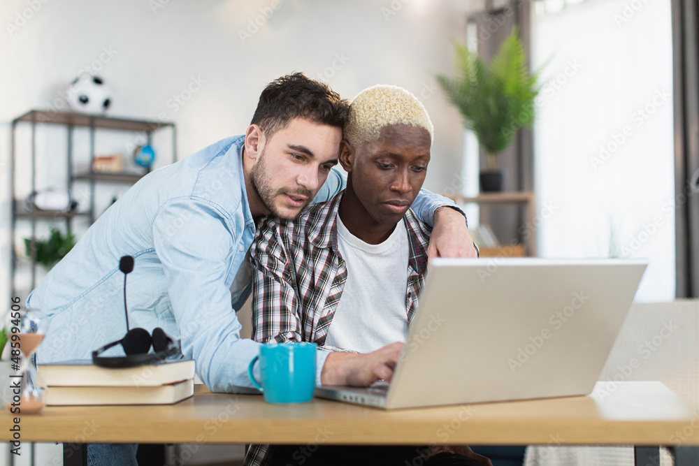 African american man sitting at desk and typing on laptop while his caucasian boyfriend standing near and embracing him. Concept of sexual orientation, technology and lifestyles. African american man sitting at desk and typing on laptop while his caucasian boyfriend standing near and embracing him. Concept of sexual orientation, technology and lifestyles.