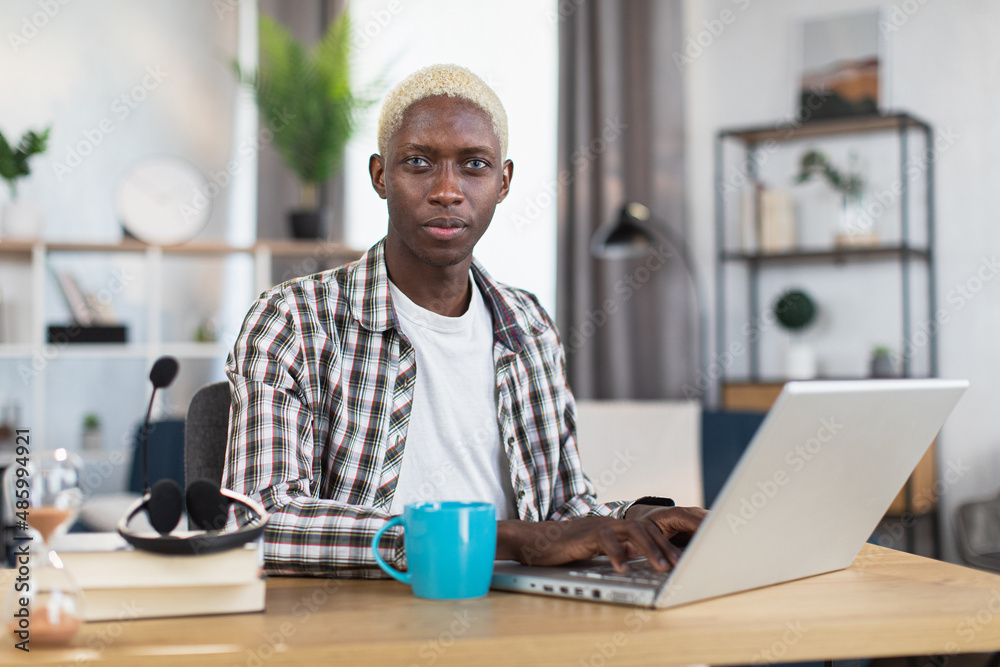 Handsome african american man with blond hair sitting desk and working on modern laptop. Male freelancer using portable computer for remote work at home. Handsome african american man with blond hair sitting desk and working on modern laptop. Male freelancer using portable computer for remote work at home.