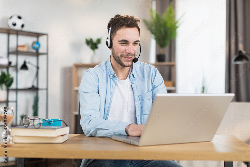 Young caucasian man in headset sitting at desk and having video call on modern laptop. Concept of gadgets, remote work and communication. Young caucasian man in headset sitting at desk and having video call on modern laptop. Concept of gadgets, remote work and communication.